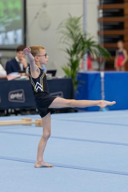 Young boy in black leotard performs a precise arabesque on the floor, leg extended high, expression focused and determined.