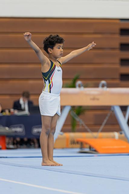 Young boy gymnast stands proudly on the floor mat, arms outstretched, presenting his routine at an indoor gymnastics meet.