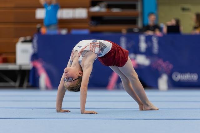 Young gymnast wearing glasses performs a bridge pose on the blue floor mat during an indoor gymnastics event.