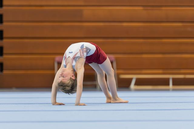 Young gymnast performs a backbend bridge on the floor mat, head lowered, body arched with focused determination.