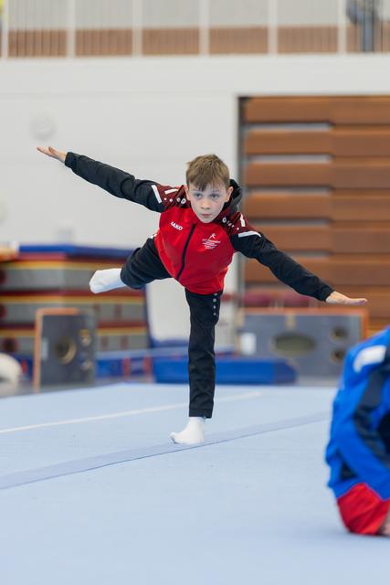 Young boy in red vest balances on one leg, arms outstretched, focused expression during floor gymnastics routine.