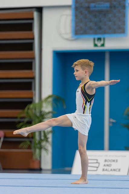 Young male gymnast balances on one leg with arms outstretched, performing a floor routine with focused concentration.