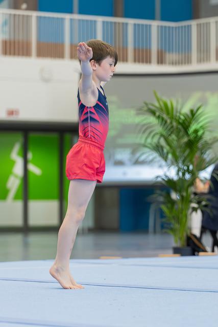 Young male gymnast stands on tiptoes on the floor mat, arm raised, focused expression during a gymnastics event.