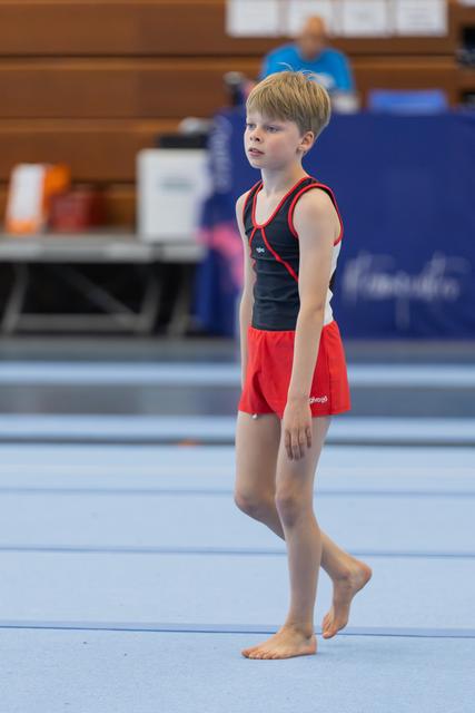Young male gymnast stands barefoot on the blue floor mat, gazing ahead with calm concentration during a gymnastics event.