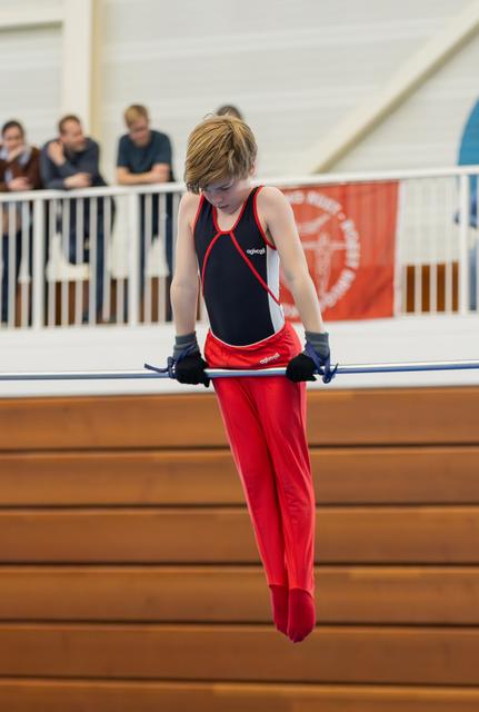 Young male gymnast grips the horizontal bar with focused intensity, wearing red trousers and a black leotard, spectators behind.