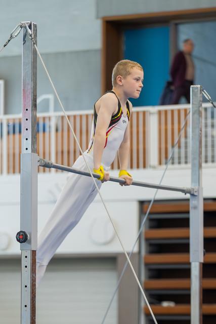 A focused young male gymnast grips the horizontal bar with yellow grips, leaning forward with intense concentration in an indoor gym.