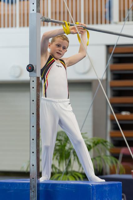 Young boy gymnast stands on a blue block, gripping the horizontal bar with focused determination, wearing white pants and a black leotard.