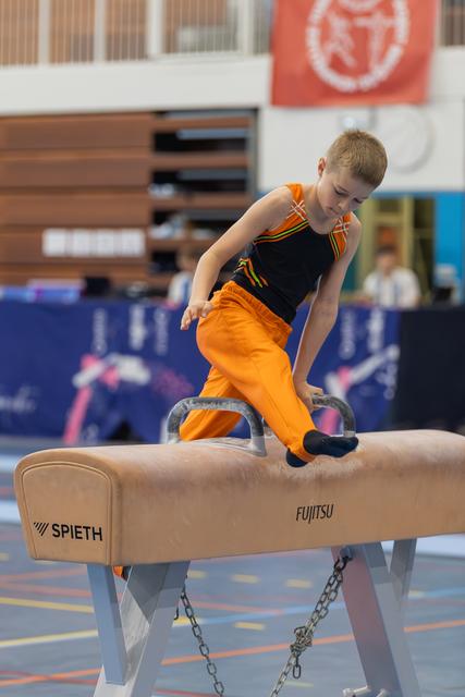 Focused young boy in orange pants performs on a Spieth pommel horse during a gymnastics competition.