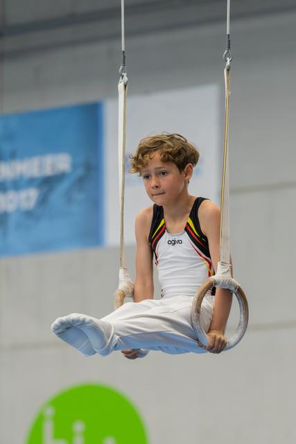 Young boy sits calmly on gymnastics rings, wearing a Belgian leotard, with a composed, attentive expression in an indoor gym.