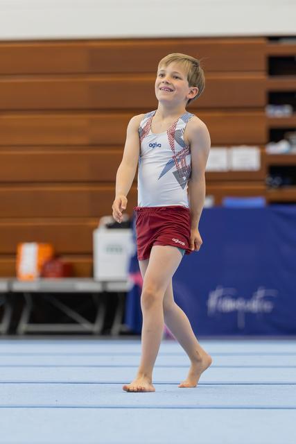 Young boy in agiva gymnastics attire walks barefoot on floor mat, smiling proudly during a gymnastics event.