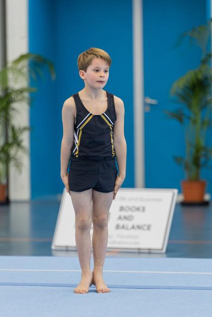Young male gymnast stands barefoot on blue mat, composed and focused, wearing black leotard with gold detailing.