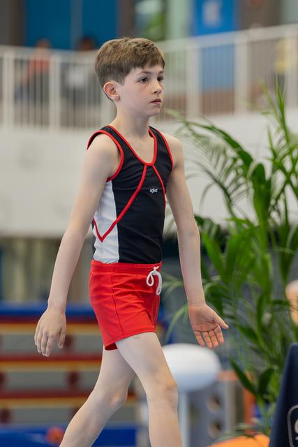 Young male gymnast walks with focused expression, wearing black and red leotard, in an indoor gymnastics venue.