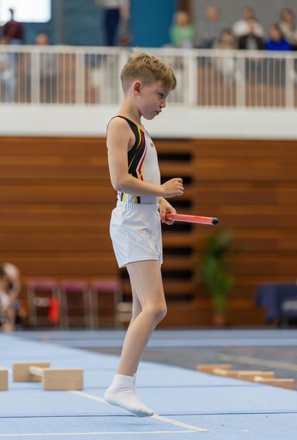 Focused young gymnast in white shorts walks across blue mat, holding a red baton during an indoor gymnastics event.