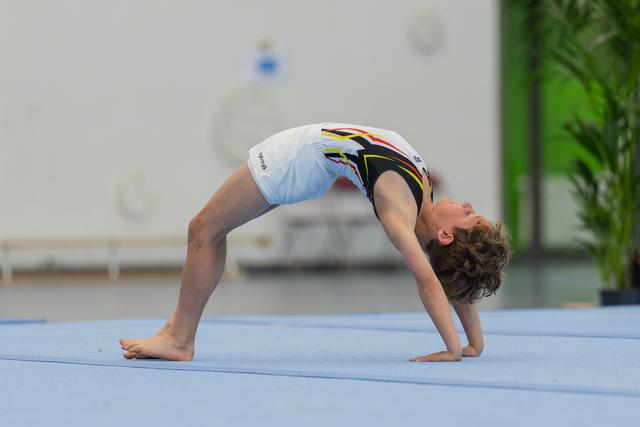 Young male gymnast performs a back bridge on blue floor mat, body arched with focused expression during floor exercise.