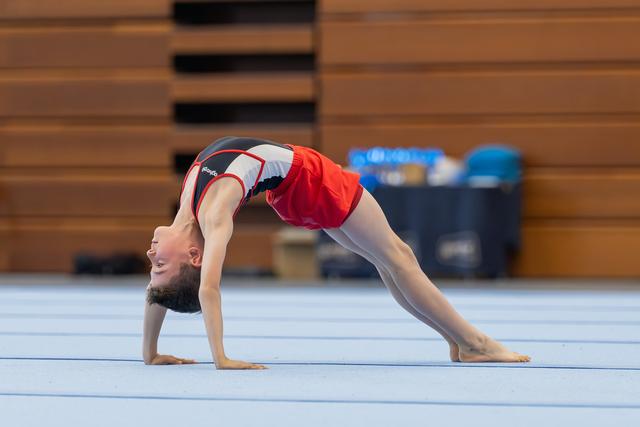 Young gymnast performs a back bridge on the floor mat, smiling upward with visible flexibility and confidence.