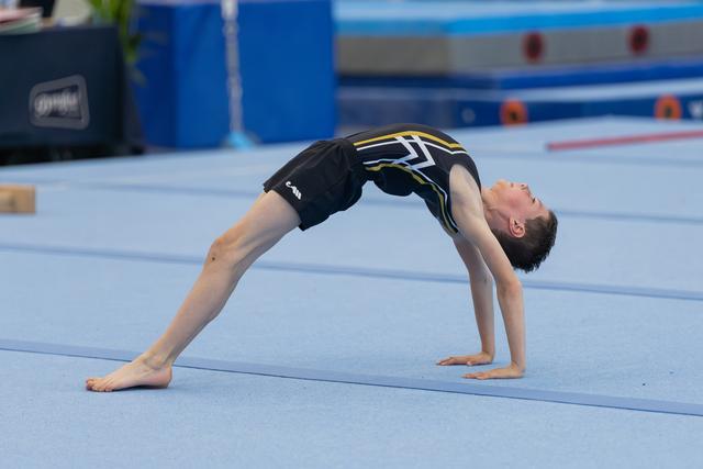 Young male gymnast holds a back bridge position on blue floor mat, body arched, focused and controlled.