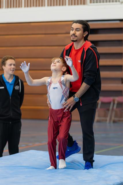 Young boy gymnast raises hands with coach steadying him from behind, both looking upward on a blue mat.