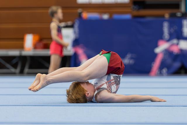 Young gymnast performs a floor routine, legs raised in a pike position, red-and-white leotard, indoor gymnastics hall.
