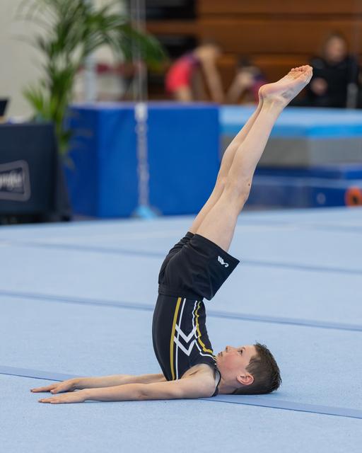 Young male gymnast performs a shoulder stand on blue floor mat, legs extended straight upward, body curved with focus.