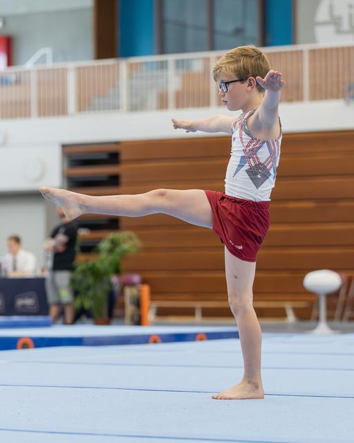 Young boy with glasses performs a precise leg kick on the floor exercise mat, arms extended, expression focused and determined.