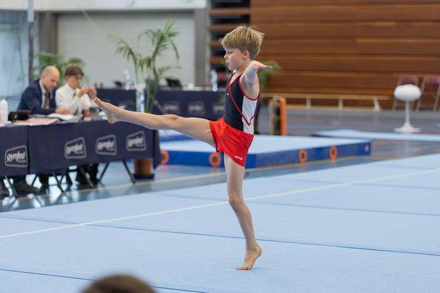 Young male gymnast performs a standing leg raise on the floor exercise mat, focused and precise, with judges scoring in background.