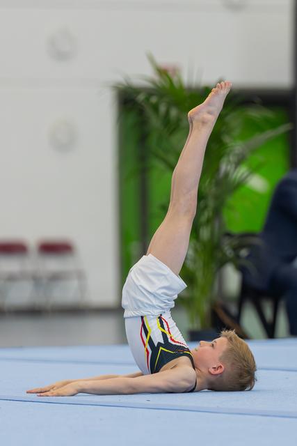 Young male gymnast lying on blue mat, legs raised vertically, performing a floor routine with focused expression.