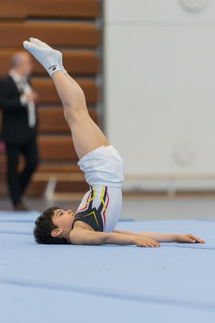Young boy performs a floor gymnastics move, lying flat with legs raised high, focused expression, on blue mat indoors.