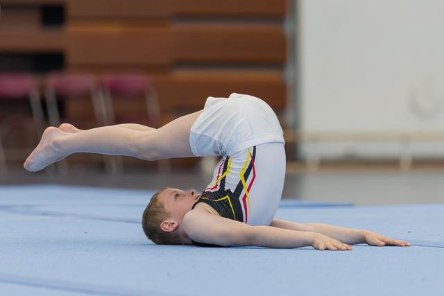 Young male gymnast performs an impressive back flexibility hold on the blue floor mat in an indoor gymnasium.