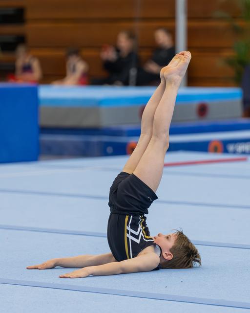Young male gymnast performs a shoulder stand on the floor exercise mat, legs pointed straight up, focused expression.