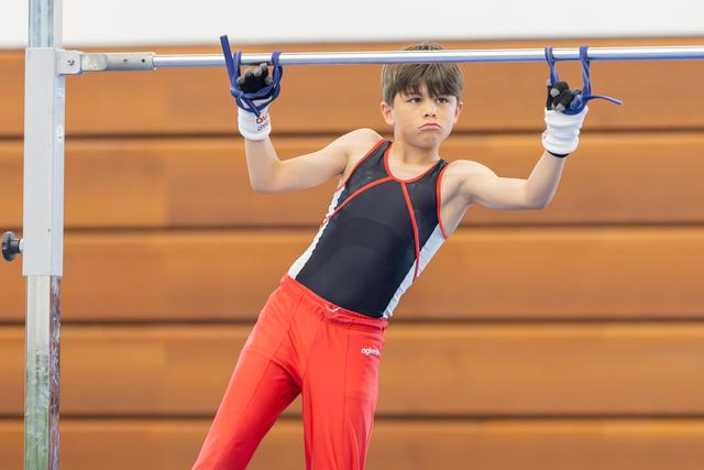 Young male gymnast grips the high bar with intense focus, wearing a black and red leotard with red pants.