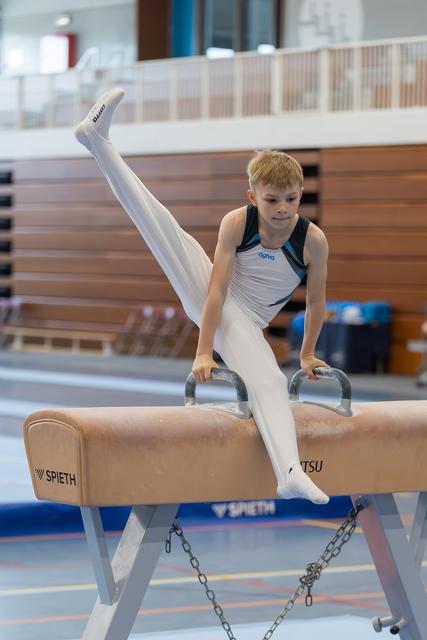 Focused young boy performs a leg split on the pommel horse, gripping the handles with determination in a bright gymnasium.