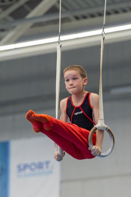 Young boy performs on gymnastic rings, legs raised, wearing red and black leotard, concentrating intently in an indoor sports hall.