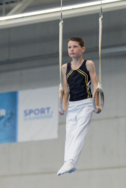 Young boy gymnast hangs focused on still rings in a sports hall, wearing black and white competition attire.