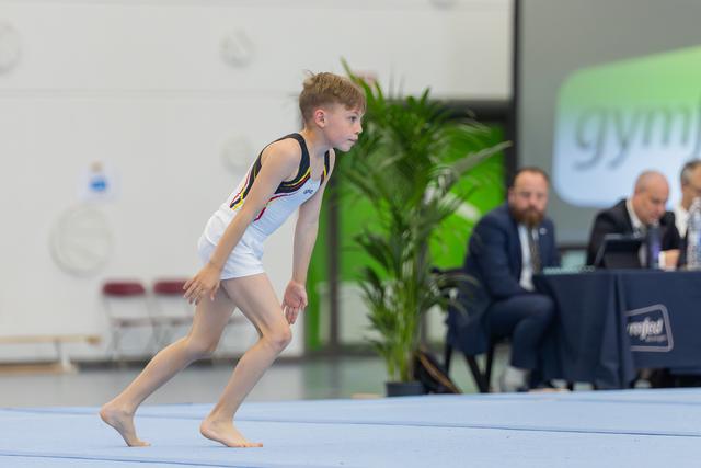 Young male gymnast crouches into a focused run-up stance on the floor exercise mat, judges seated behind.