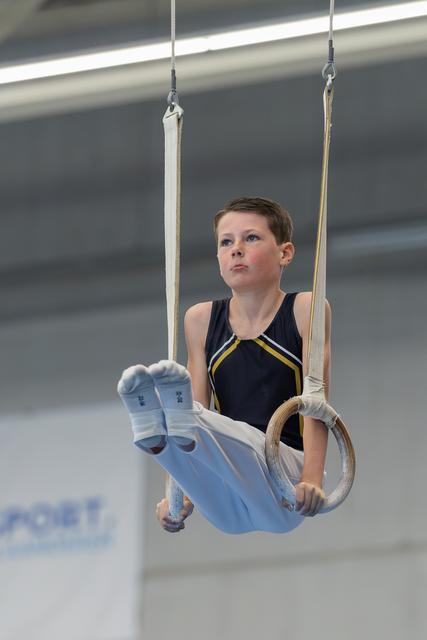 Young boy gymnast performs on still rings, legs raised, wearing chalk-dusted grips, expression focused and determined.