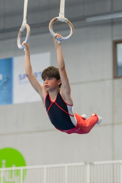 Focused young male gymnast hangs from still rings in a navy and red leotard during an indoor gymnastics competition.