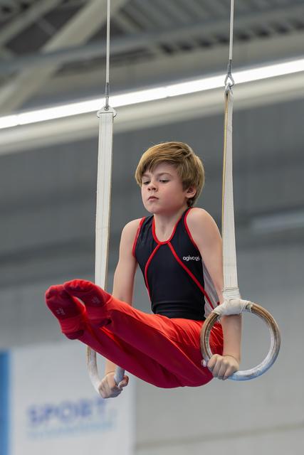 Focused young boy grips still rings mid-routine, legs tucked, wearing black and red gymnastic suit indoors.