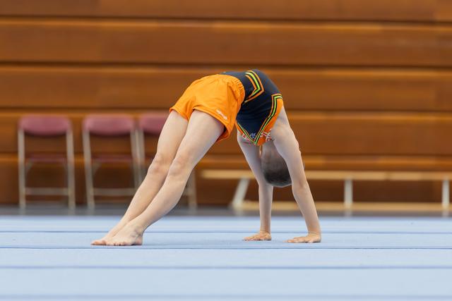 Young gymnast bent forward in a wide-legged stretch on the blue floor mat in a gymnasium.