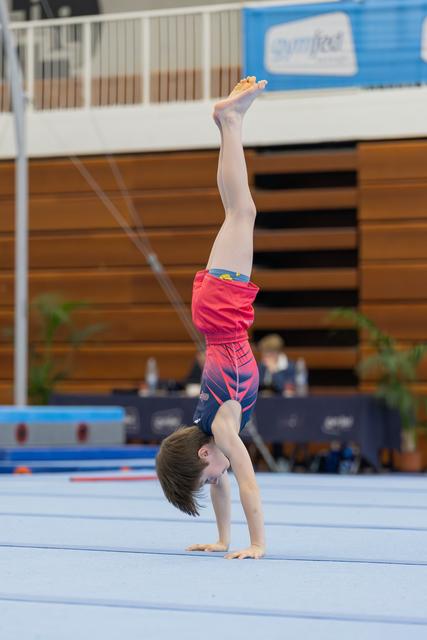 Young boy performs a straight handstand on the floor exercise mat during a gymnastics competition, legs extended upward.