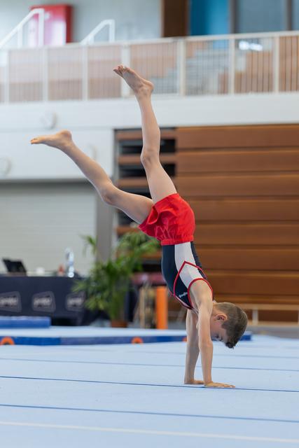Young male gymnast performs a handstand with legs split apart on the floor exercise mat in an indoor gymnastics hall.
