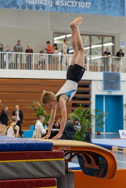 Young male gymnast performs a handstand on the vault, legs straight up, concentrated expression in a busy sports hall.