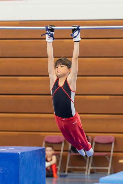Young male gymnast hangs from the high bar with intense focus, wearing red and black gymnastics attire in an indoor gym.