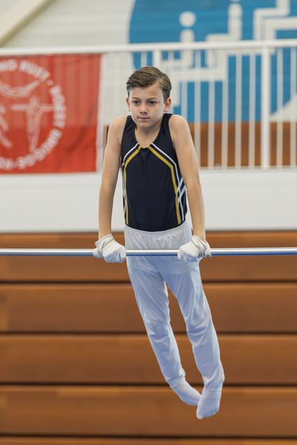 Young male gymnast hangs from the high bar with steady grip and calm, focused expression in a gymnasium.