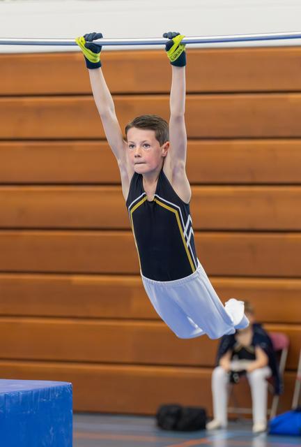 Determined young boy hangs from horizontal bar in black gymnastics uniform, arms fully extended, expression intense and focused.