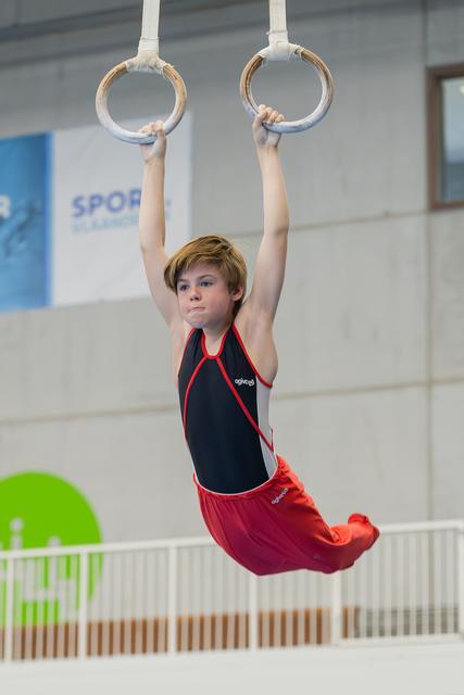 Young boy hangs from gymnastic rings with intense focus, body suspended mid-air in an indoor gymnastics hall.