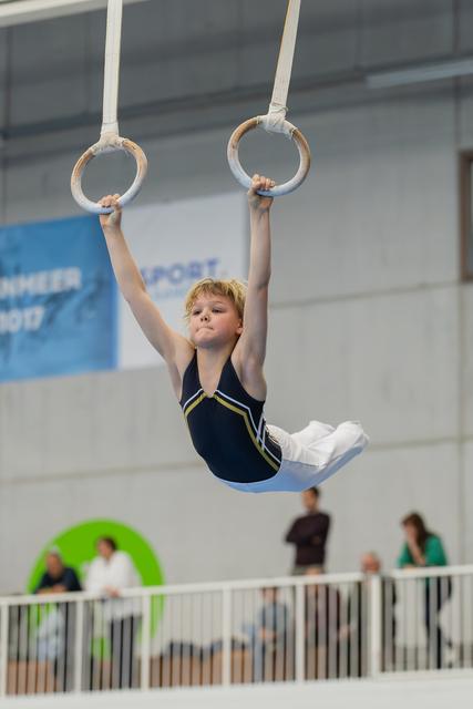 Young boy grips gymnastics rings mid-routine, legs tucked, face set with intense concentration in an indoor sports hall.