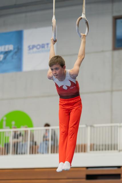 Young male gymnast hangs from still rings with focused expression, wearing red and white leotard in indoor gymnasium.