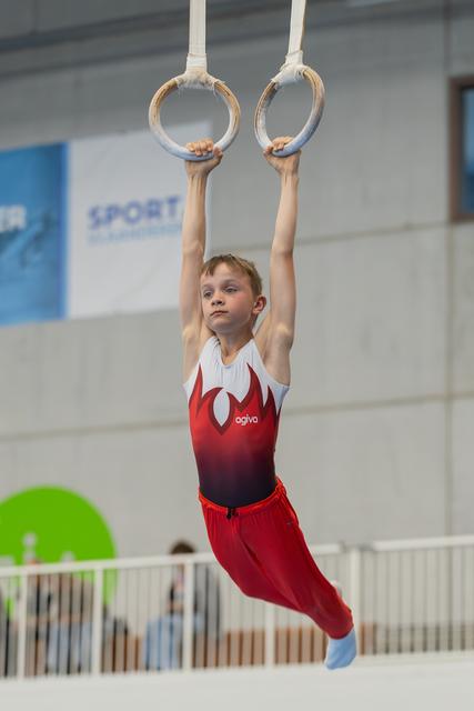 Young boy in red Agiva leotard hangs from gymnastics rings with focused expression in a sports hall.