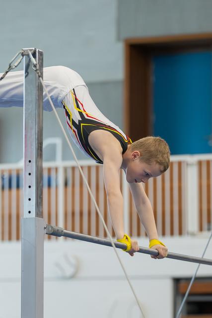 Young boy gymnast grips the high bar with yellow hand guards, body horizontal, concentrating during a gymnastics event.
