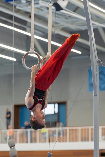 Young male gymnast hangs inverted on still rings, legs raised, wearing red and black, performing in an indoor gymnasium.
